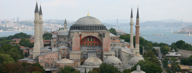 <p><strong>Ayasofya Cami</strong></p>

<p>Fatih Sultan Mehmed’in İstanbul’u fethettiğinde ilk cuma namazını kılmaya layık gördüğü Ayasofya ihtişamı ile tüm dünyanın gözdelerinden biri olarak şanslı İstanbul’un ve tarihi yarımadanın da gözdesidir.</p>

<p>537 yılında tamamlanan ve İmparator Justinianos tarafından yaptırılan Ayasofya 916 yıl Bizans’ta kilise olarak kullanıldıktan sonra Fetih Sultan Mehmed’in İstanbul’u 1453’te fethinden itibaren 482 yıl cami olarak hizmet vermiştir.<br />
Yapı içerisinde H.z. İsa, Meryem Ana, Konstantin, Büyük Meleklerden Cebrail gibi figürler altın, gümüş ve mermer mozaiklerle bezenmiştir.</p>

<p>Ayasofya İstanbul’un fethinden sonra Fatih Sultan Mehmed tarafından camiye çevrilirken ilk minaresini de Fatih döneminde eklenmiştir. Daha sonra 2. Bayezid dönemlerinlde de eklenen minareler olmuştur.<br />
Osmanlı döneminde Ayasofya içerisine Minber, Mihrab, Vaaz kürsüsü eklenerek Cami hüviyetine bürünmüştür.<br />
Ayasofya Mimar Sinan’ın yaptığı  destekler sayesinde günümüzdeki gibi sağlam ayakta kalabilmiştir.</p>

<p>Günümüzde Müze olarak hizmet veren Ayasofya Müzesi bahçesi içerisinde birçok pahitaht mensubu ve padişah türbeleri de yer almaktadır. İçerisindeki kapıları, mezar taşları ve Osmanlı döneminde eklenen yapıları ile Ayasofya hala eski ihtişamını koruyor.</p>

