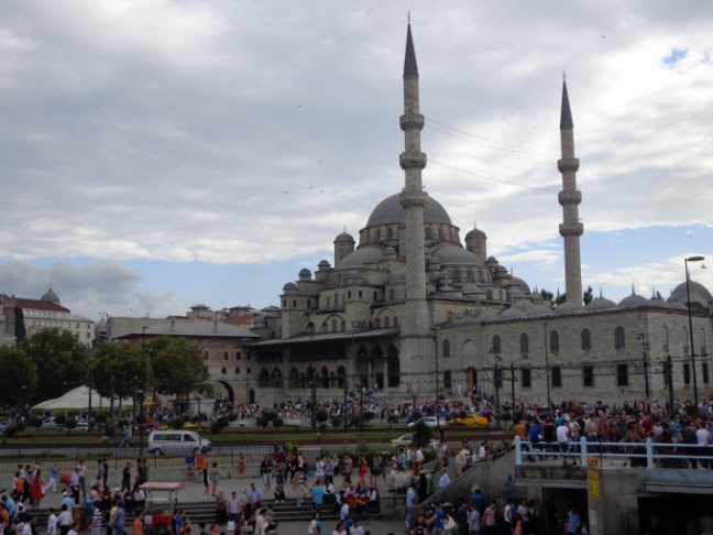 <p><strong>Eminönü Yeni Camii</strong></p>

<p>İstanbul’un en kalabalık yerlerinden biri olan Eminönü meydanında bulunan Eminönü Yeni Cami, namı diğer Valide Sultan Cami önünden belkide defalarca geçtiğiniz bir yapı. Osmanlı Devleti döneminde yapımı en uzun süren yapı olarak da tarihe geçmiş bir yapıdır.</p>

<p>Öyle ki yapının başlangıcından bitimine Osmanlı Devleti Tahtından 3. Murat, 3.Mehmet, 1. Ahmet, 1. Mustafa (2 Kez tahta çıktı bu sürede), 2. Osman(Genç Osman), 4. Murat, 1. İbrahim, 4. Mehmet olmak üzere 8 Osmanlı Padişahı geçmiştir.</p>

<p>1589 yılında 3. Murat’ın karısı Safiye Sultan adına yapımına başlanan caminin yapımı o kadar uzun sürmüş ki Safiye Sultan adına yaptırılan camiyi görememiş. Cami yapımı önce Davut Ağa’nın Veba salgınında vefat etmesi ve sonrasında ise 3.Mehmet ve Safiye Sultan’ın vefatı ile bir süre durdurulduktan sonra 1661 yılında bu kez bir başka Valide Sultan, 4. Mehmet’in annesi Turhan Sultan tarafından yapımı yeniden başlatılmıştır. Neyse ki bu başlangıç sonrası inşaat kısa sürmüş ve 2. yılın sonunda 1663 yılında cami bir cuma namazında ibadete açılmıştır.</p>

<p>Cami yapımına başlayan Mimar Sinan’ın talebesi Davut ağa, Dalgıç Mehmet Ağa’ya devrettiği inşaatı yarım yüzyıl sonra tamamlamak Mustafa Ağa’ya nasip olmuştur.</p>

<p>Cami Külliyesinde; Mısır Çarşısı, Türbe ve Hünkar Kasrı’ndan oluşan yapılar yer alırken, Yeni Cami (Valide Sultan Cami) türbesinde; Hatice Sultan ile birlikte  2. Mustafa, 1. Mahmut, 3. Ahmet ve 4.Mehmet’in sandukaları bulunur.</p>

<p>Eminönü Yeni Cami (Valide Sultan Cami) külliyesinde yer alan Mısır Çarşısı günümüzde de tüm ihtişamı ve kalabalığı ile işlemeye devam etmektedir.</p>

<p>Bugün Eminönü Yeni Cami deniz tarafındaki kapısından baktığınızda karşınızda tüm ihtişamı ile Galata Kulesi ve Köprüsü yer alırken, bugün deniz ile Külliye arasında kalan geniş alan cami temelleri atıldığında tamamen denizle kaplıymış.</p>

<p>Eminönü Yeni Cami ve Külliyesi etrafındaki manzarası, külliye içerisindeki çeşitlilik dolayısıyla turistler ve fotoğrafçılar açısından eşsiz bir yapı olarak İstanbul’un göbeğinde yer almaktadır.  Bu kalabalığın içinde normal vakit namazlarında yaklaşık 1500, Cuma namazlarında ise 7000-8000 kadar bir cemaate ibadet etme şansı vermektedir.</p>
